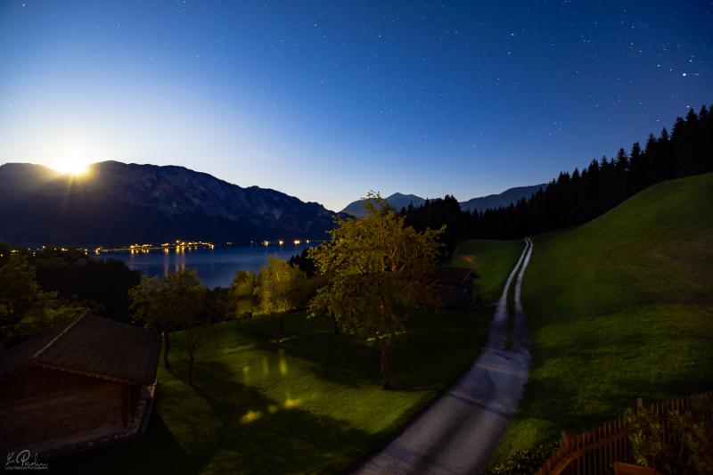 Long exposure photo of the moon rising in a blue sky at night. In the background alps and the Attersee, Austria